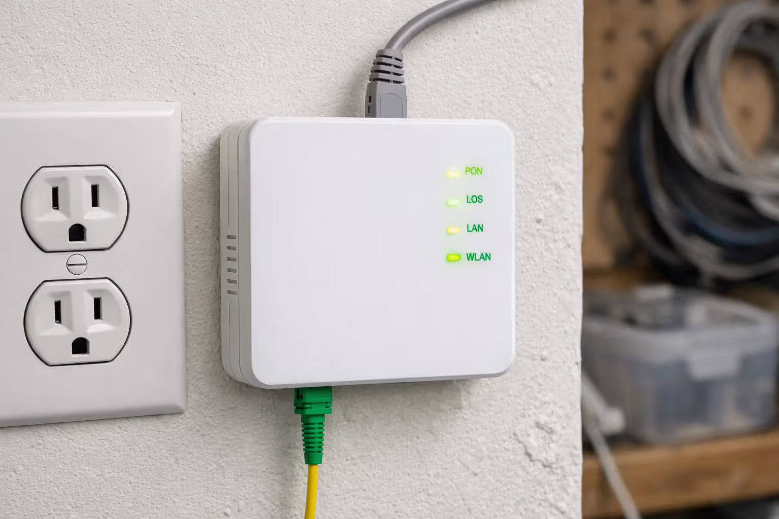 Close-up of a white Optical Network Terminal (ONT) mounted on a wall with a fiber optic cable plugged in at the bottom and an ethernet cable connected at the top, green LED status lights glowing